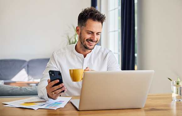 man sitting at a desk using a laptop holding a coffee cup and smartphone surrounded by papers promoting productivity tips for 12 tasks