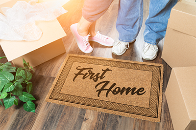 couple standing on doormat that says First Home surrounded by moving boxes representing new beginnings and homeownership steps